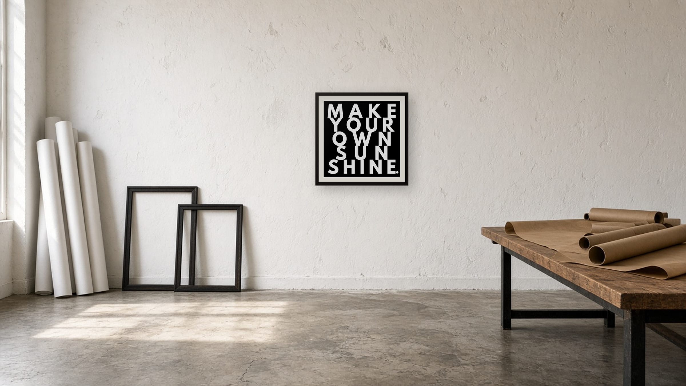 Make Your Own Sunshine print, white on black, framed on a white studio wall near a worktable with kraft paper and shipping tubes.